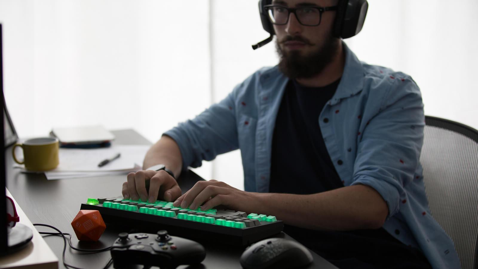 A man wearing a gaming headset sits at a desk with a gaming keyboard, an Xbox controller, and a Dungeons & Dragons die.