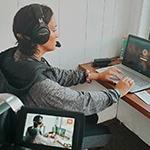 毕业生悉尼菲利普斯, 一个深棕色头发的女人，穿着灰色连帽运动衫，戴着黑色耳罩式耳机, 坐在写字台前的笔记本电脑前的记者包厢里俯瞰着足球场上穿着制服的运动员.